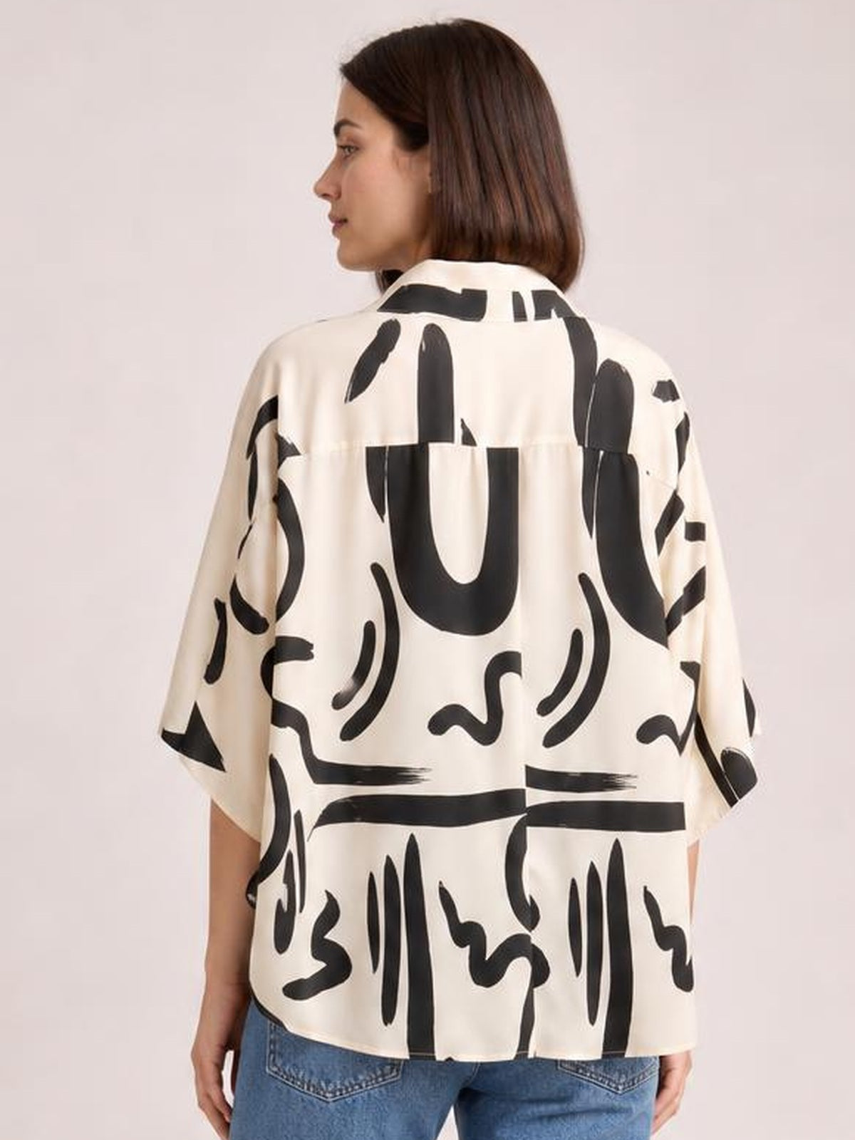 A model turned in profile shows the back of a cream printed blouse with bold black abstract patterns, a notched collar, half sleeves, and a relaxed silhouette.