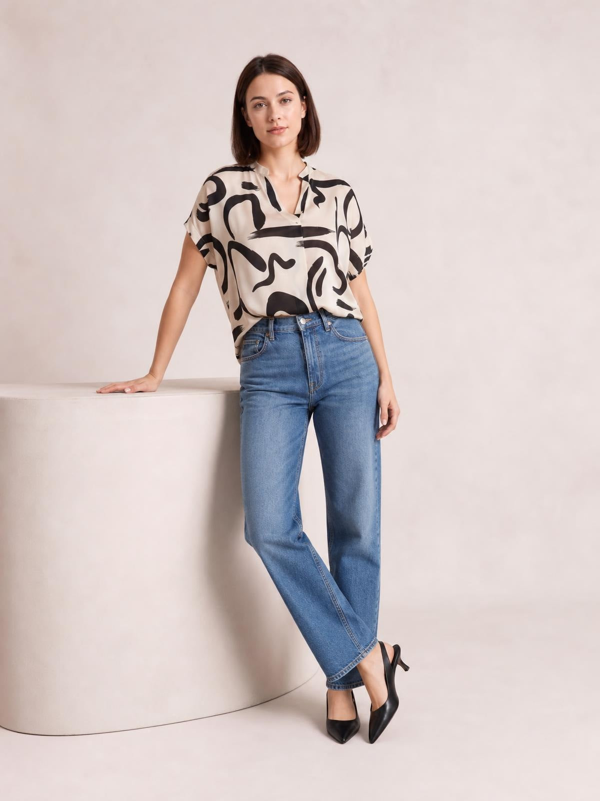 Model wears an opaque printed blouse with a notched neckline, short sleeves, relaxed silhouette, and cream fabric patterned with bold black abstract shapes.