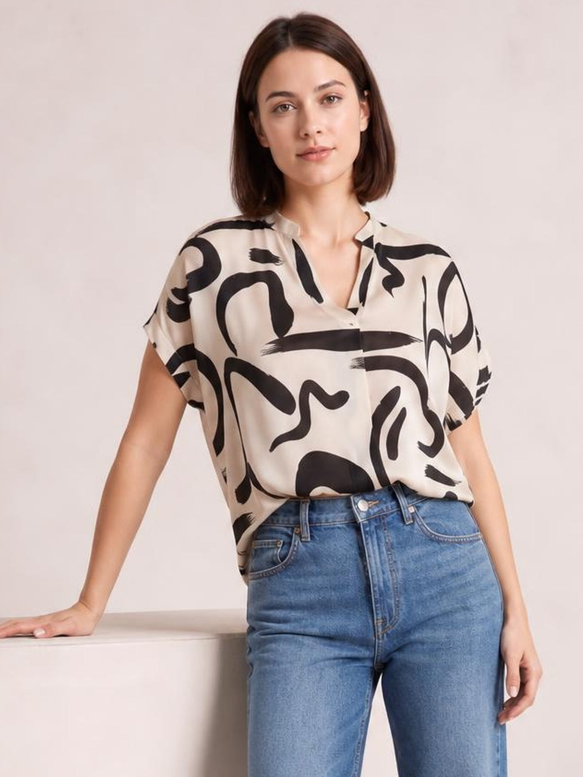 A model wears a cream printed blouse with bold black abstract shapes, a notched neckline, half sleeves, and a relaxed silhouette, paired with jeans.