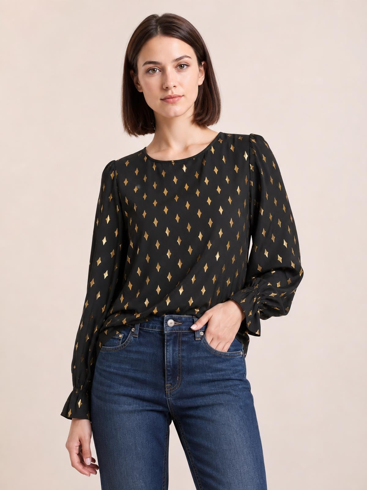 A model wears a black printed blouse with gold diamond motifs, round neckline, long puff sleeves, and gathered cuffs, posing with one hand in her jeans pocket.