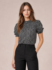 Model wearing a black printed round neck blouse with short puff sleeves, an opaque relaxed silhouette, and one hand in her trouser pocket.