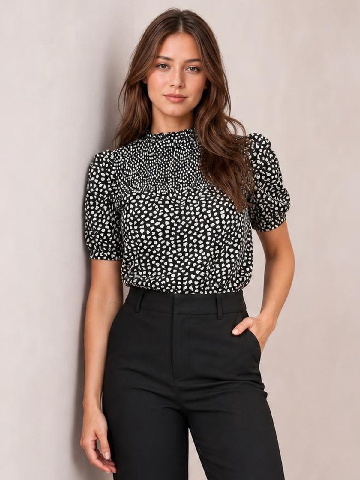 Model wearing a black printed round neck blouse with short puff sleeves and a relaxed long silhouette, paired with black trousers and one hand in a pocket.