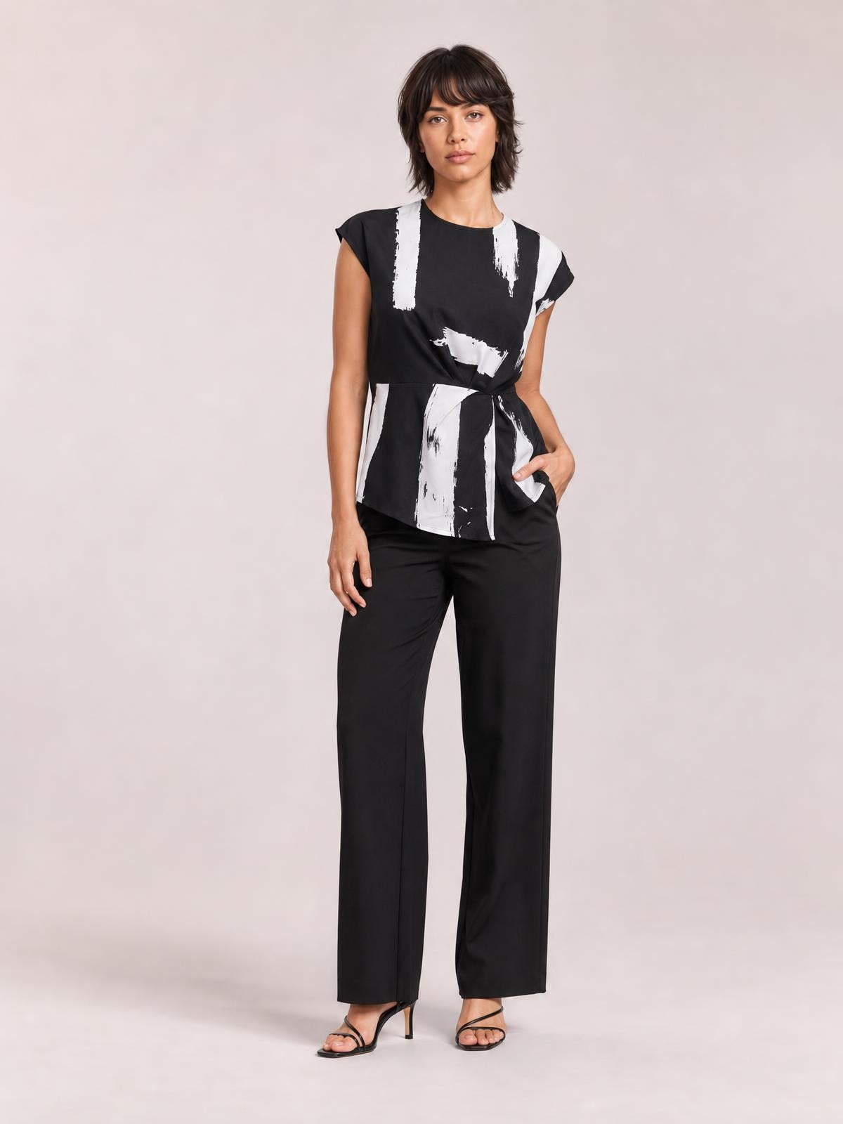 A model stands with one hand in her pocket wearing a black-and-white printed round-neck blouse with short sleeves, a waist seam, and a slightly flared long silhouette.