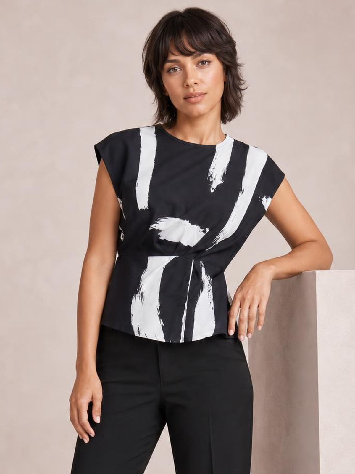 Model poses with one hand resting on a ledge, wearing a black printed blouse with a round neck, short regular sleeves, and a long, relaxed silhouette.