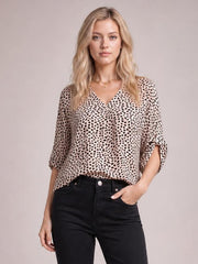 Model poses in a beige and black printed V-neck blouse with a relaxed silhouette and three-quarter roll-tab sleeves, paired with black jeans.