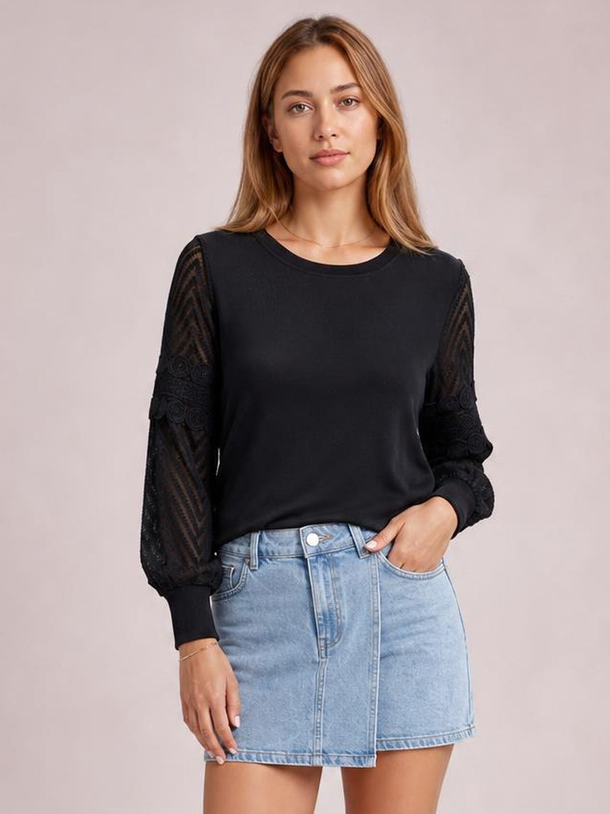 A woman models a solid black round-neck blouse with a regular-length silhouette, long patterned lace sleeves, ribbed cuffs, and a relaxed hand-in-pocket pose.