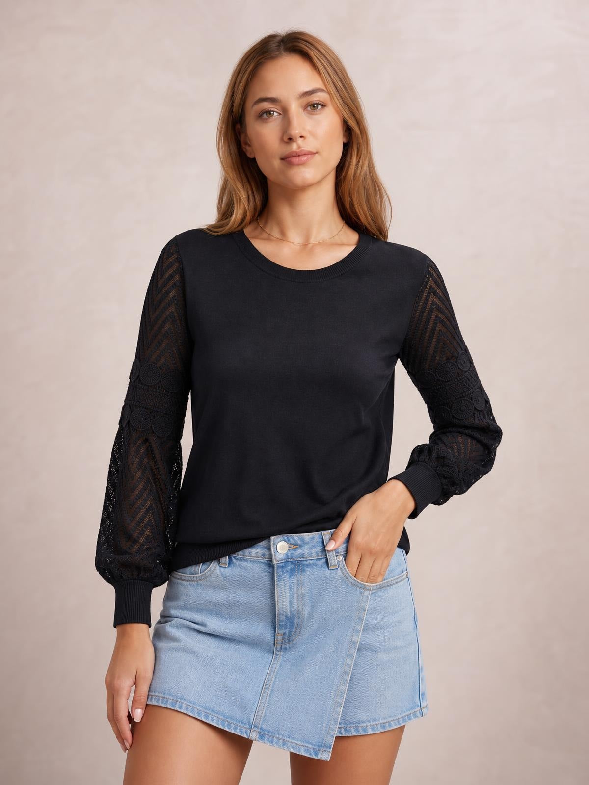 A model poses in a black round neck blouse with a regular silhouette, long patterned sheer sleeves, ribbed cuffs and hem, and one hand in her pocket.