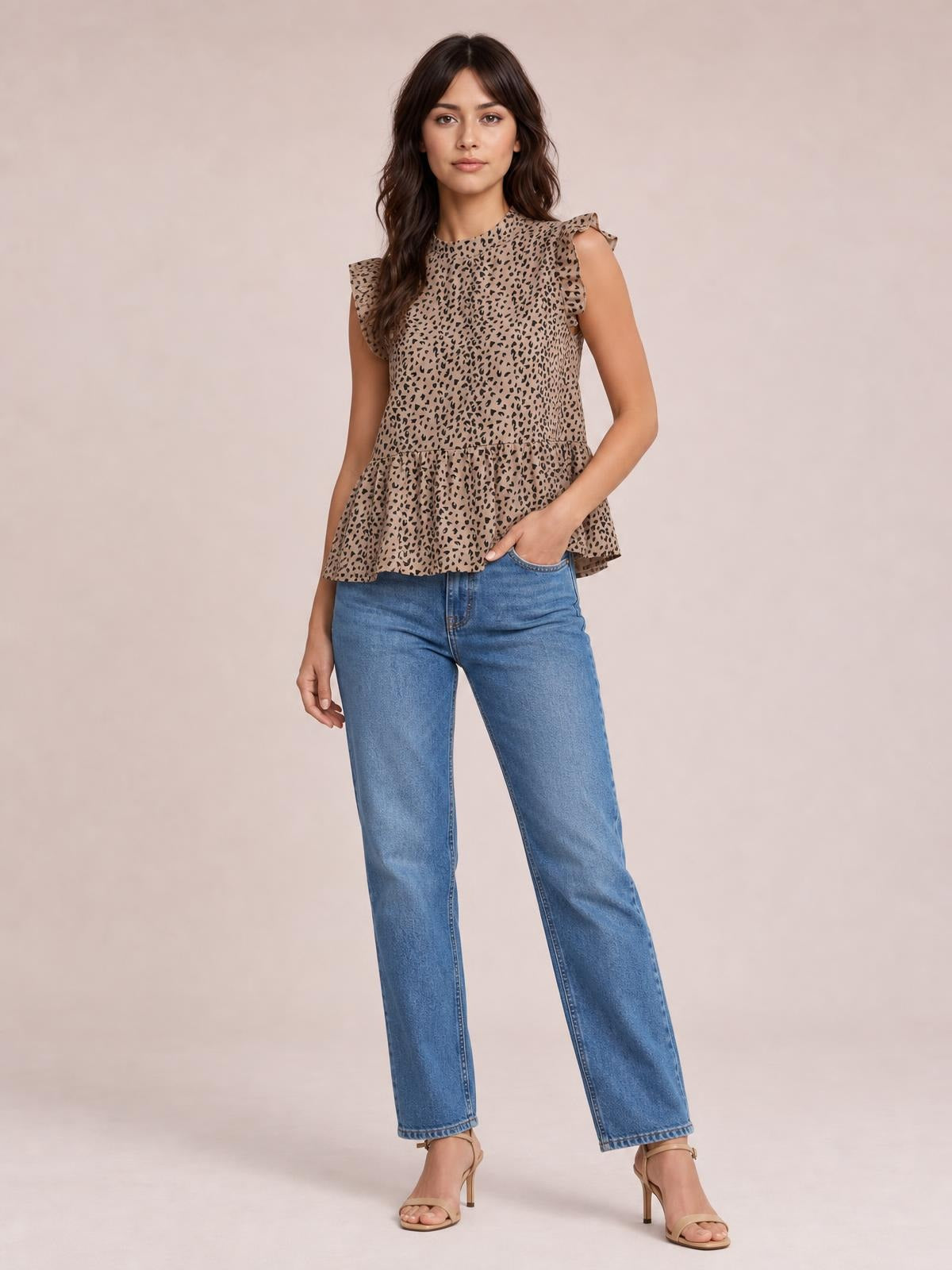 Model wears a tan and black printed mock neck blouse with ruffled cap sleeves and a flared peplum silhouette, posing with one hand in her jeans pocket.