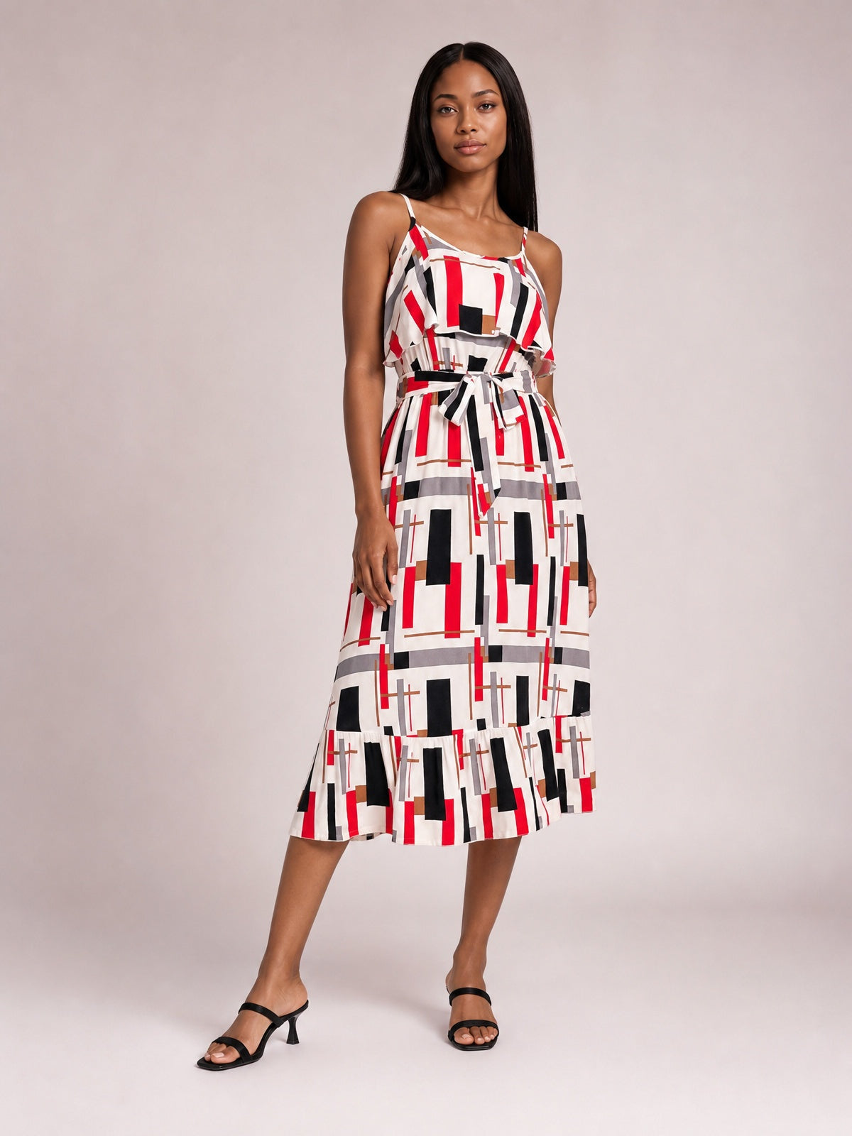 Model poses in a geometric multi-color printed midi dress with slender straps, ruffled bodice overlay, self-tie waist, and flared hem.