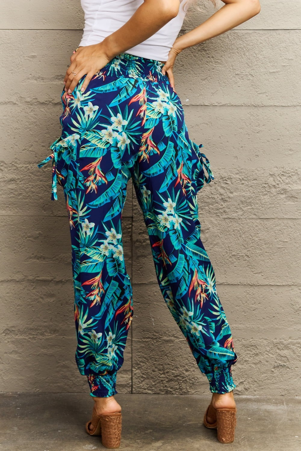 Model poses with hands on hips in long navy smocked pants featuring a colorful tropical plant print, relaxed wide-leg silhouette, and elastic ankle cuffs.