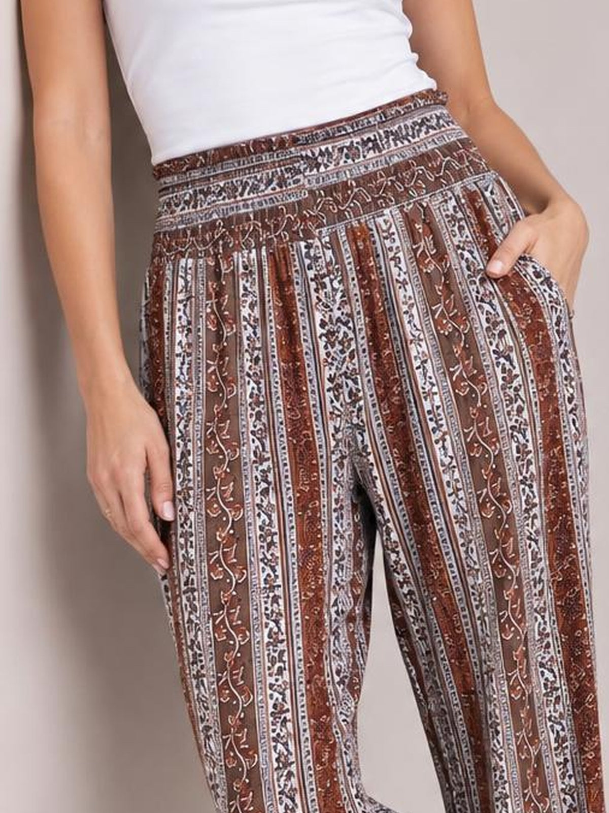 Model poses with one hand in the pocket of brown and cream printed high-waist pants featuring a smocked waistband and straight-leg silhouette.