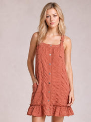Model wearing a rust textured mini dress with a square neckline, wide straps, front buttons, side pockets, relaxed silhouette, and ruffled hem.