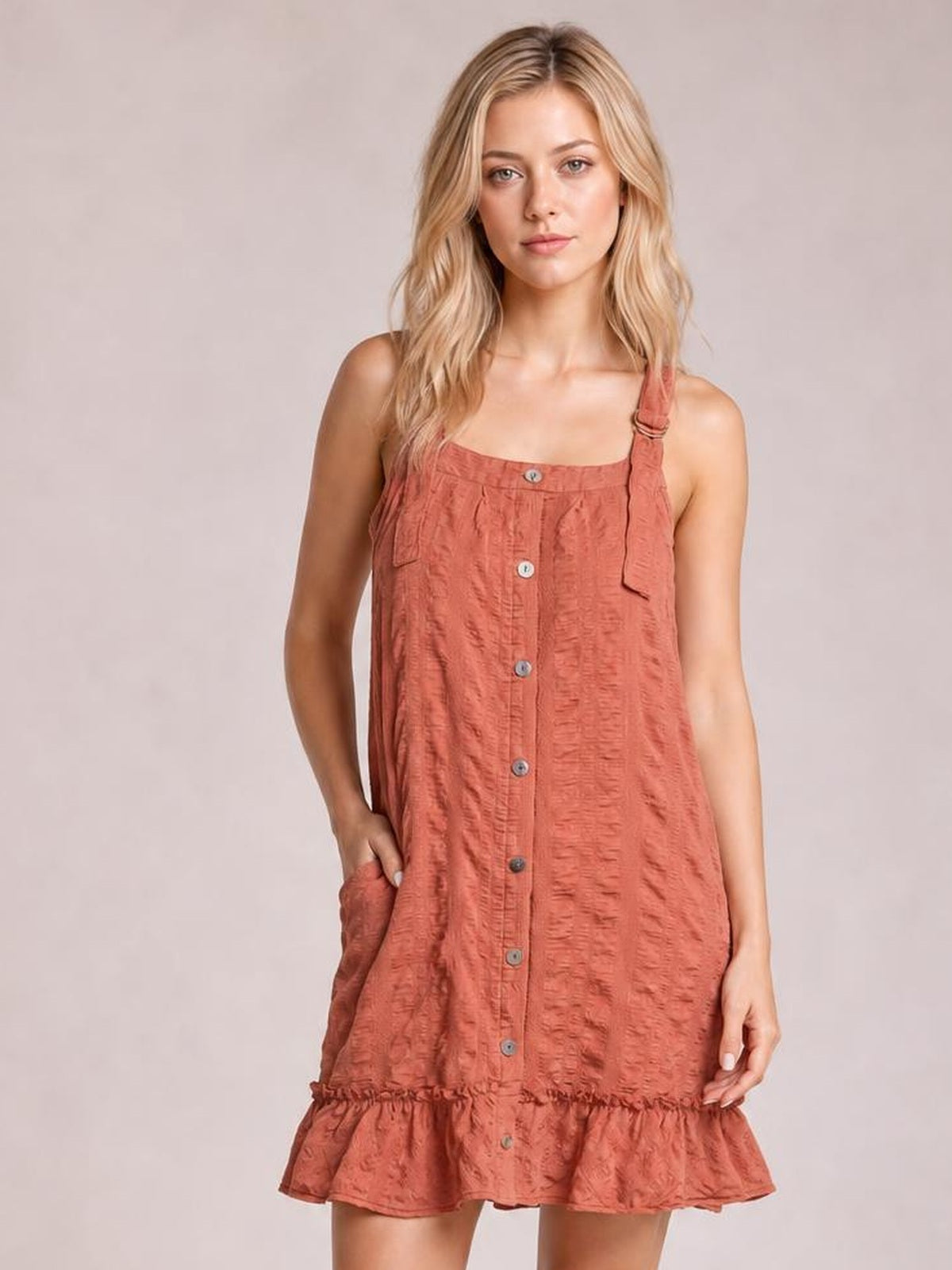 Model wearing a rust textured mini dress with a square neckline, wide straps, front buttons, side pockets, relaxed silhouette, and ruffled hem.