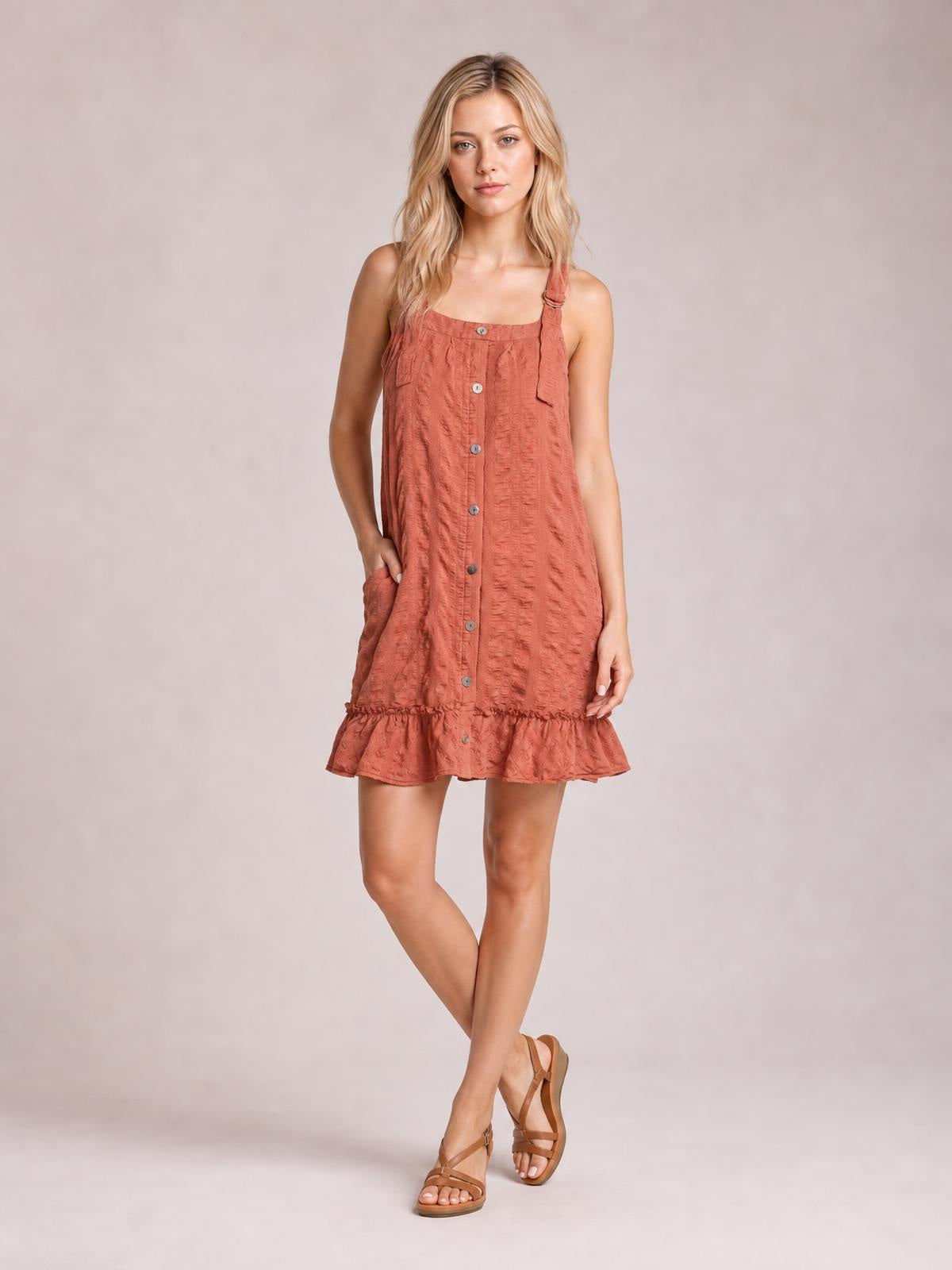 A model poses in a textured coral mini dress with a square neck, wide straps, button-front construction, side pocket, relaxed silhouette, and ruffled hem.