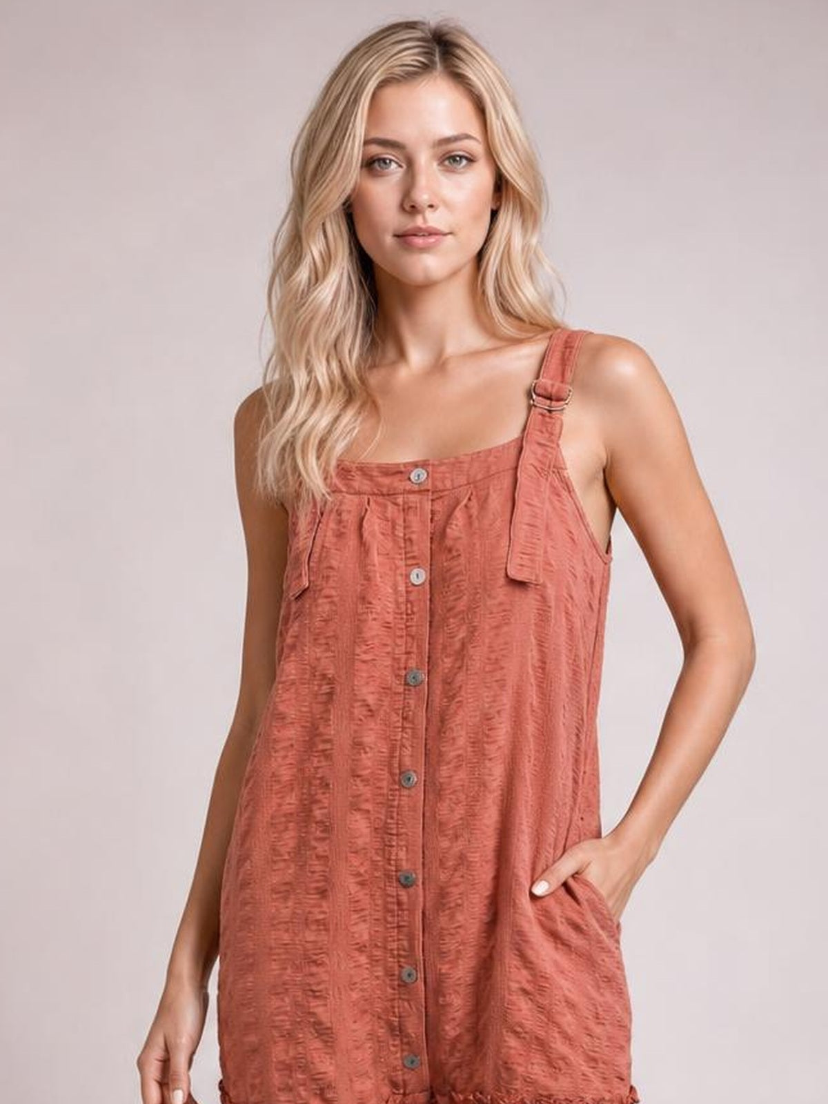 Model wears a coral textured mini dress with a square neckline, wide adjustable straps, front buttons, and side pockets, posing with one hand tucked into a pocket.