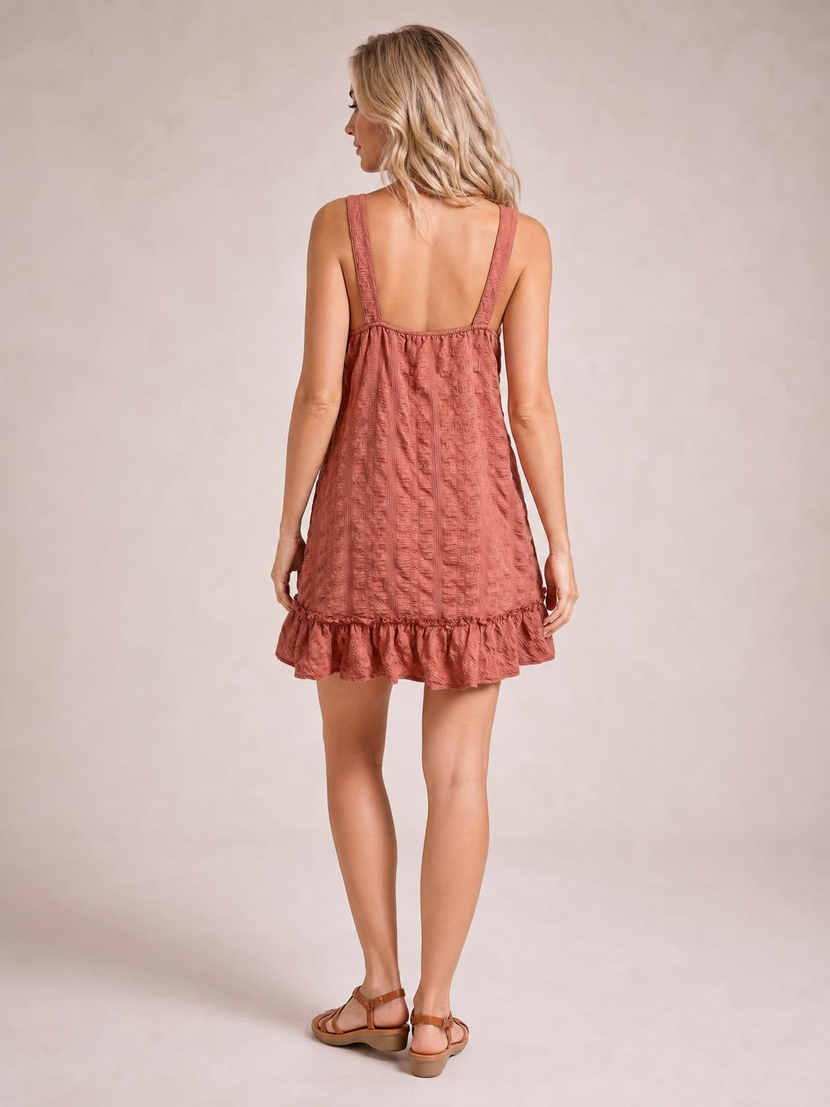 Model shown from the back in a rose-brown textured mini dress with wide straps, a relaxed A-line silhouette, and a ruffled hem.