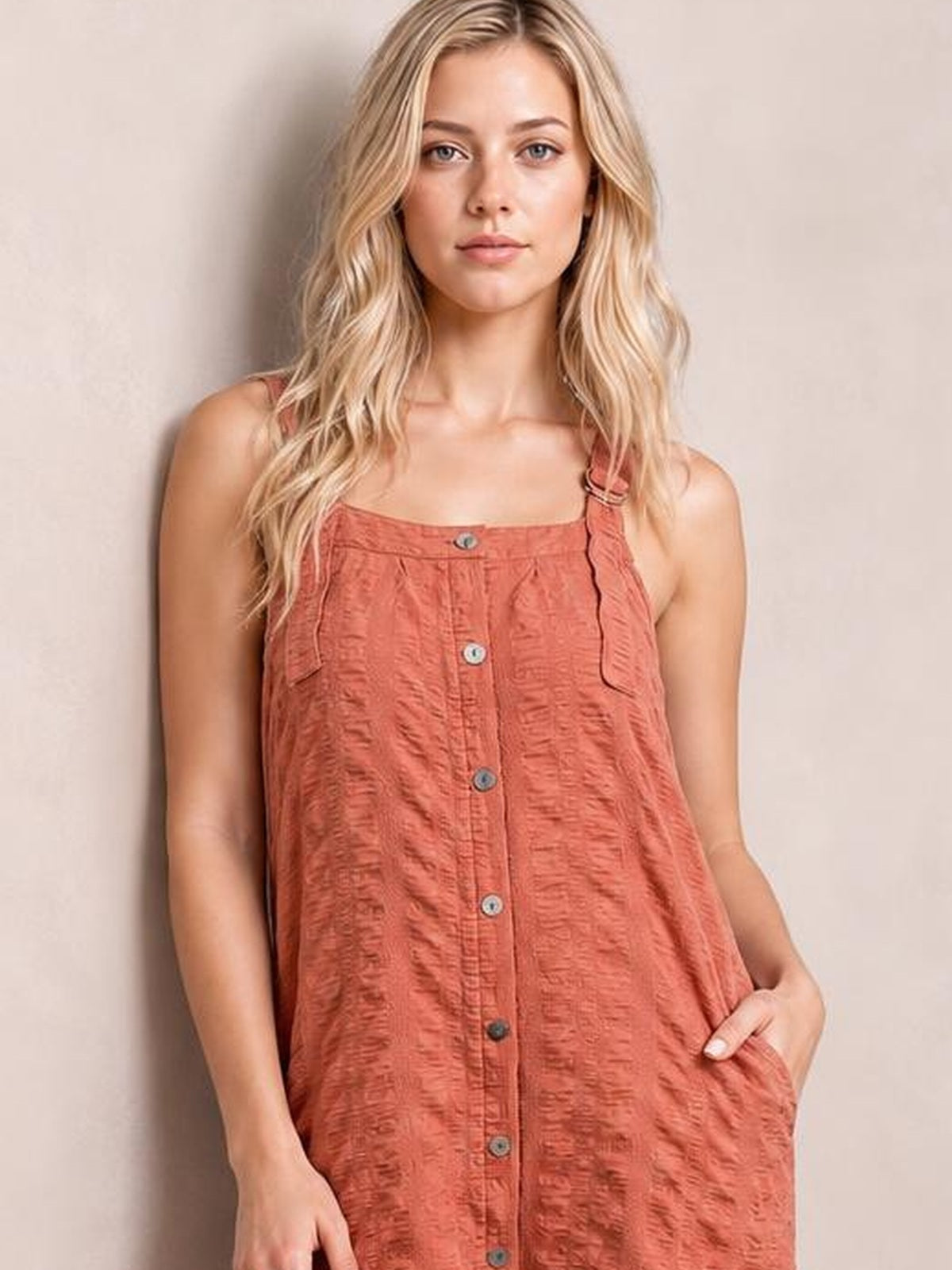 A model wears a rust textured mini dress with a square neckline, wide straps, button-front construction, relaxed silhouette, and practical side pockets.