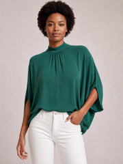 Model stands in a teal tie back mock neck blouse with ruched neckline, split three-quarter sleeves, and a relaxed, flowing silhouette.