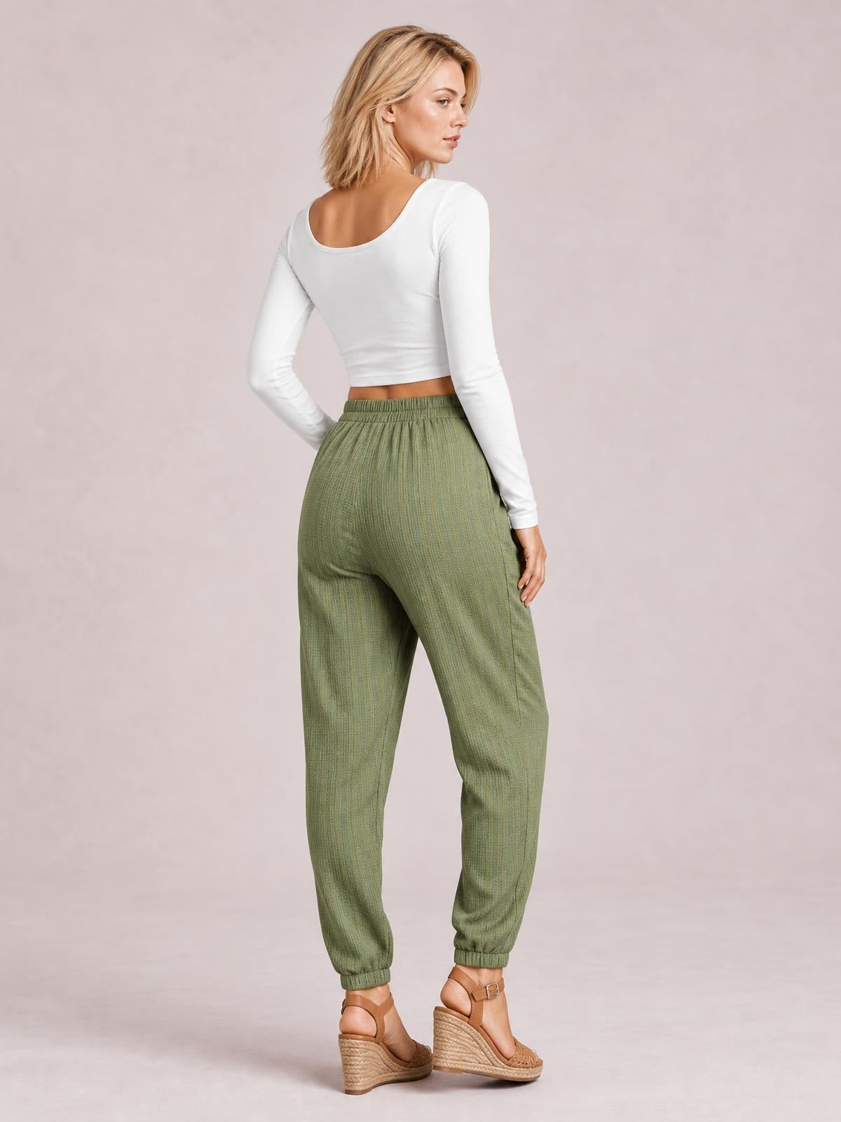 A model poses turned away in olive-green solid long pants with a gathered elastic waist, relaxed tapered legs, elastic cuffs, and one hand in a pocket.