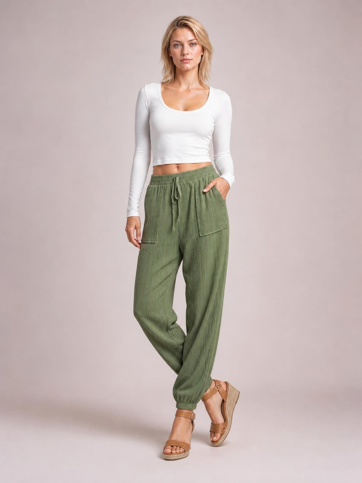 A model poses in olive green tie waist long pants with a relaxed tapered silhouette, side pockets, and gathered ankle cuffs.