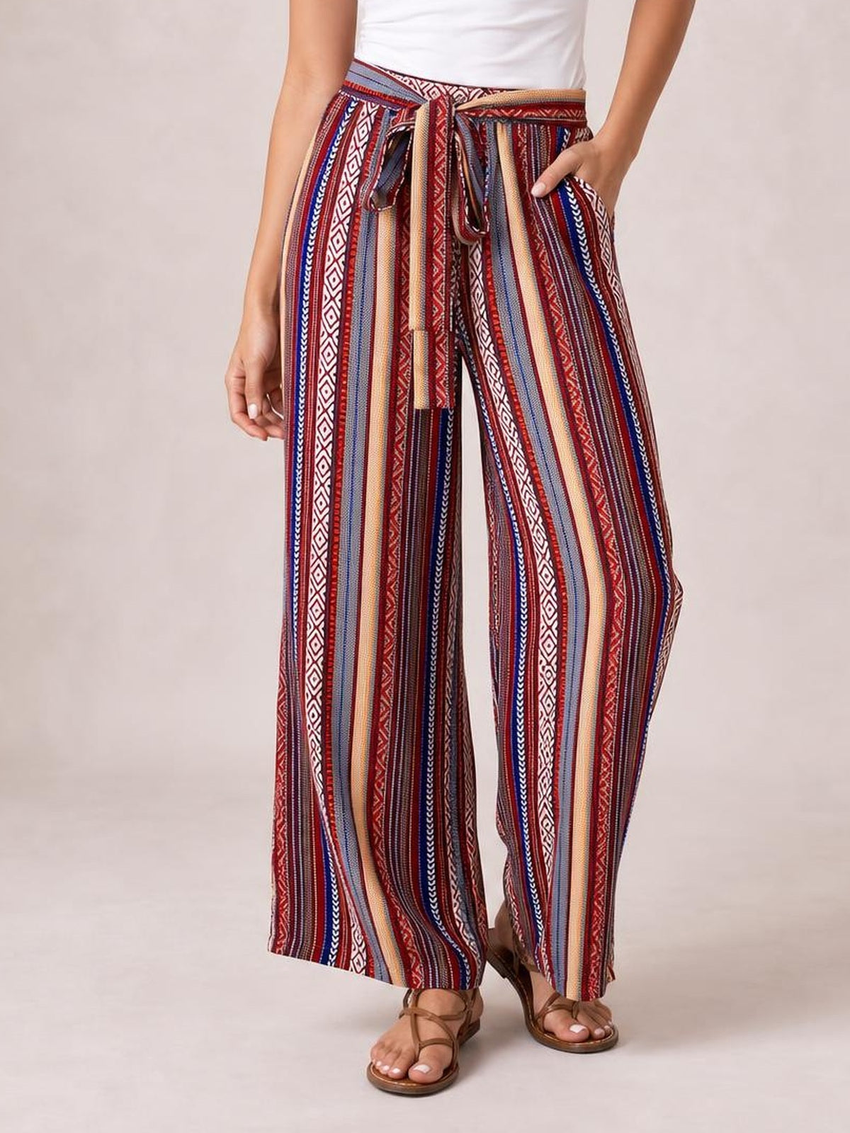 A model stands in tied geometric wide-leg pants with red, blue, cream, and black vertical stripes, a flowy silhouette, pockets, and one hand tucked in a pocket.