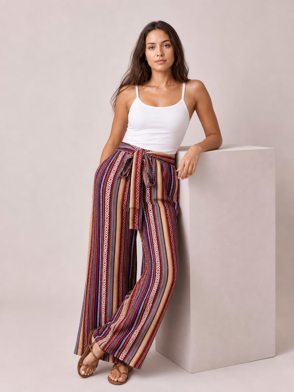 Model leans against a block wearing colorful geometric striped tied wide-leg pants with a flowy silhouette, high waist, and visible pockets.