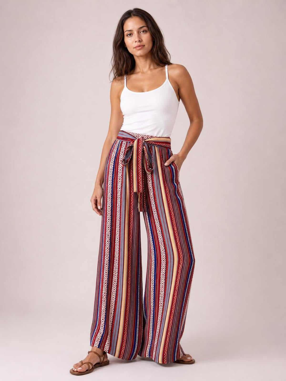 Model wears multicolored geometric wide-leg pants with vertical stripes, a tied waist, flowing silhouette, and side pockets, posing with one hand tucked inside.