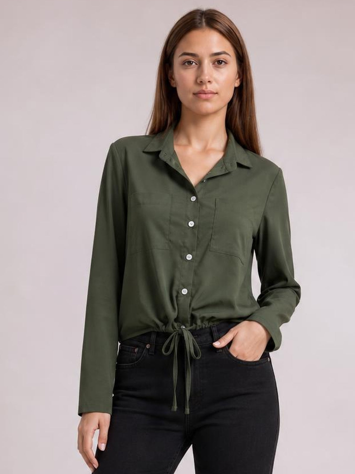 Model wearing an olive green long sleeve button-down shirt with a collared neckline, chest pocket, straight silhouette, and tied hem, standing with one hand in a pocket.