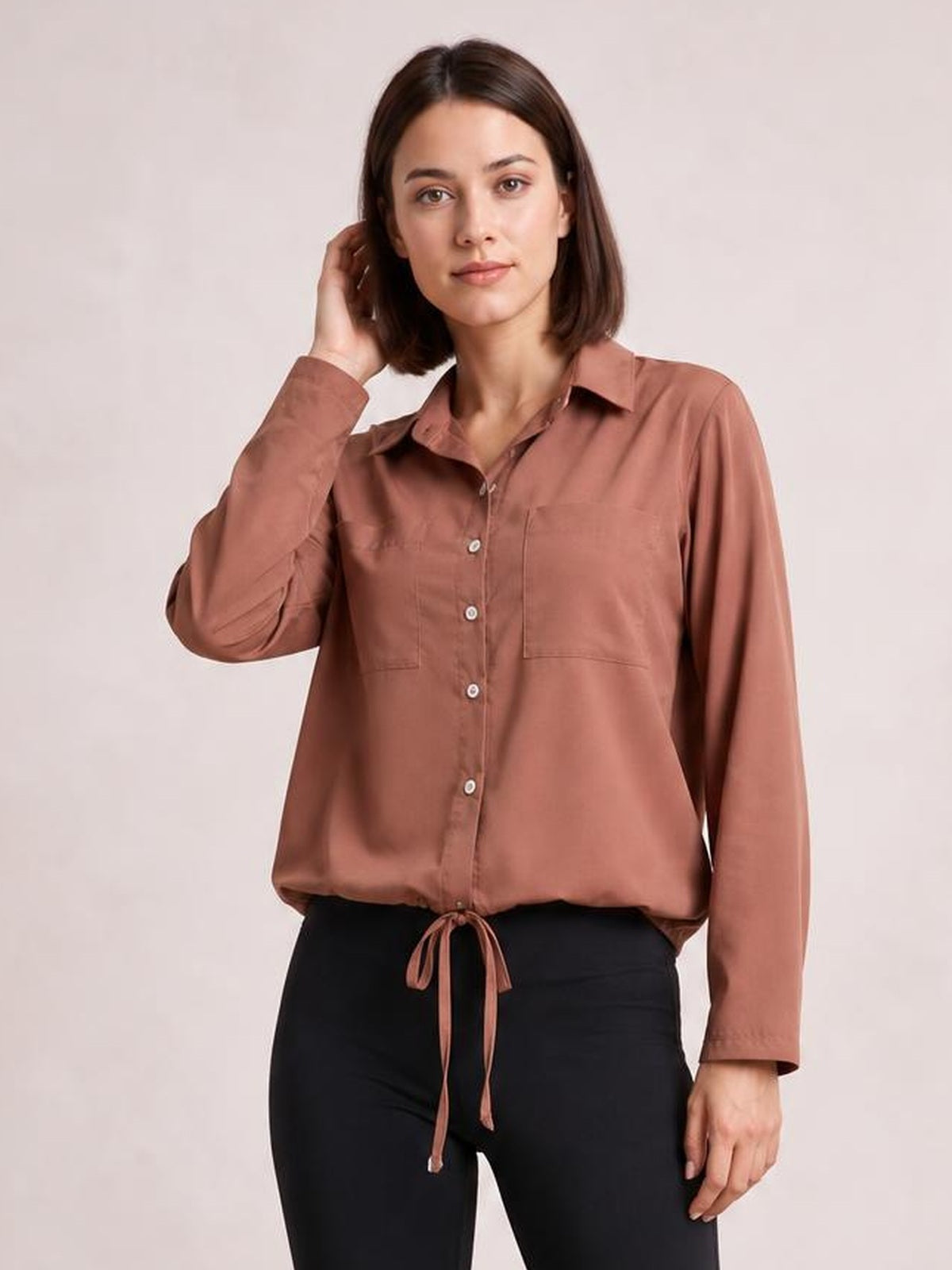 Model wearing a rust-brown tied hem button-down long sleeve shirt with a collared neckline, front buttons, chest pockets, and a relaxed silhouette.