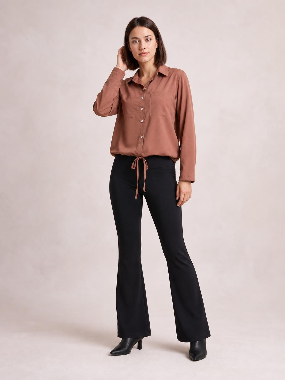 A model poses in a muted brown button-down long sleeve shirt with a pointed collar, chest pockets, and a tied hem creating a softly cropped silhouette.