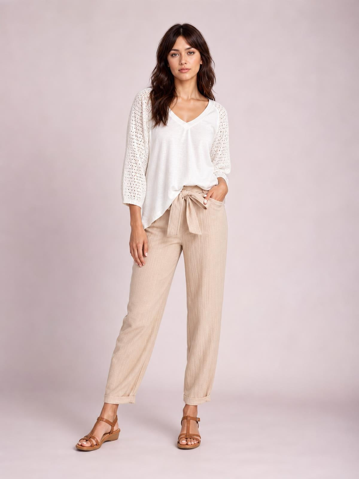 A model poses in beige tied high-waist pants with a relaxed straight-leg silhouette, visible side pockets, and cuffed hems.