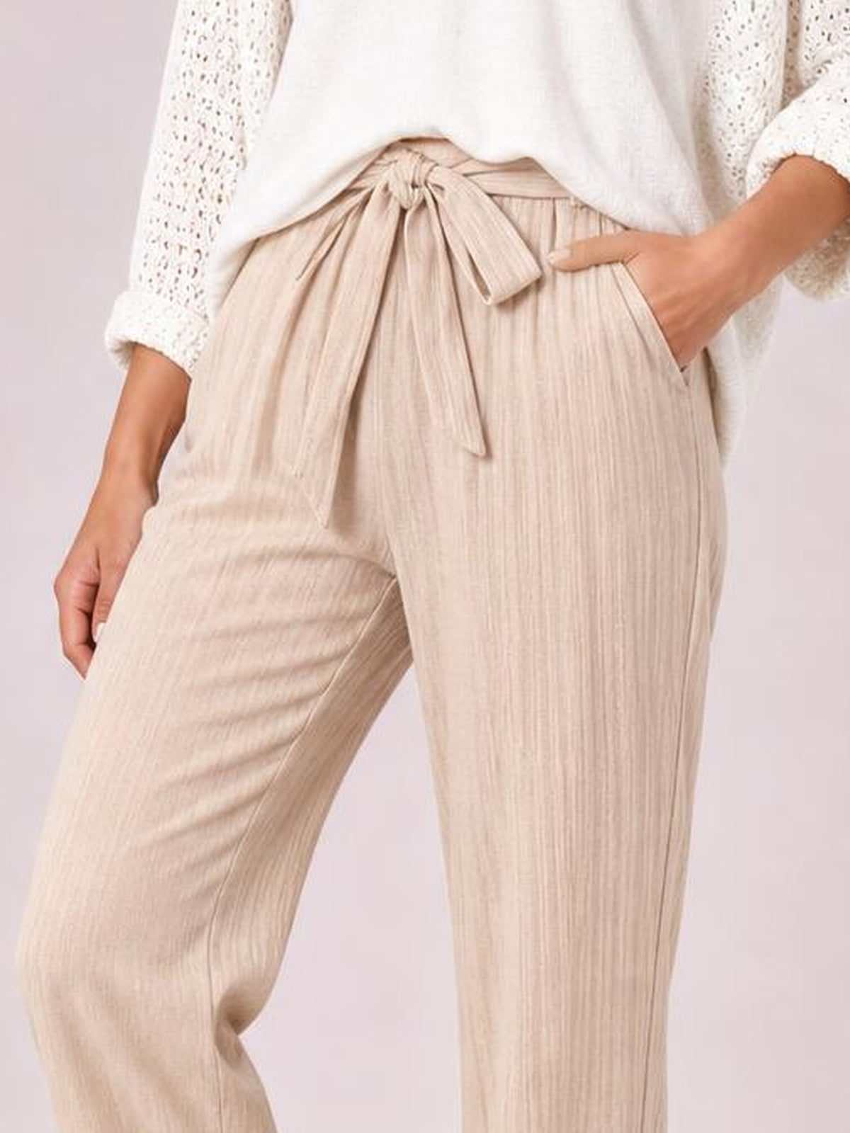 Model poses in beige high-waist pants with a tied waistband, button details, side pockets, and a relaxed straight-leg silhouette.