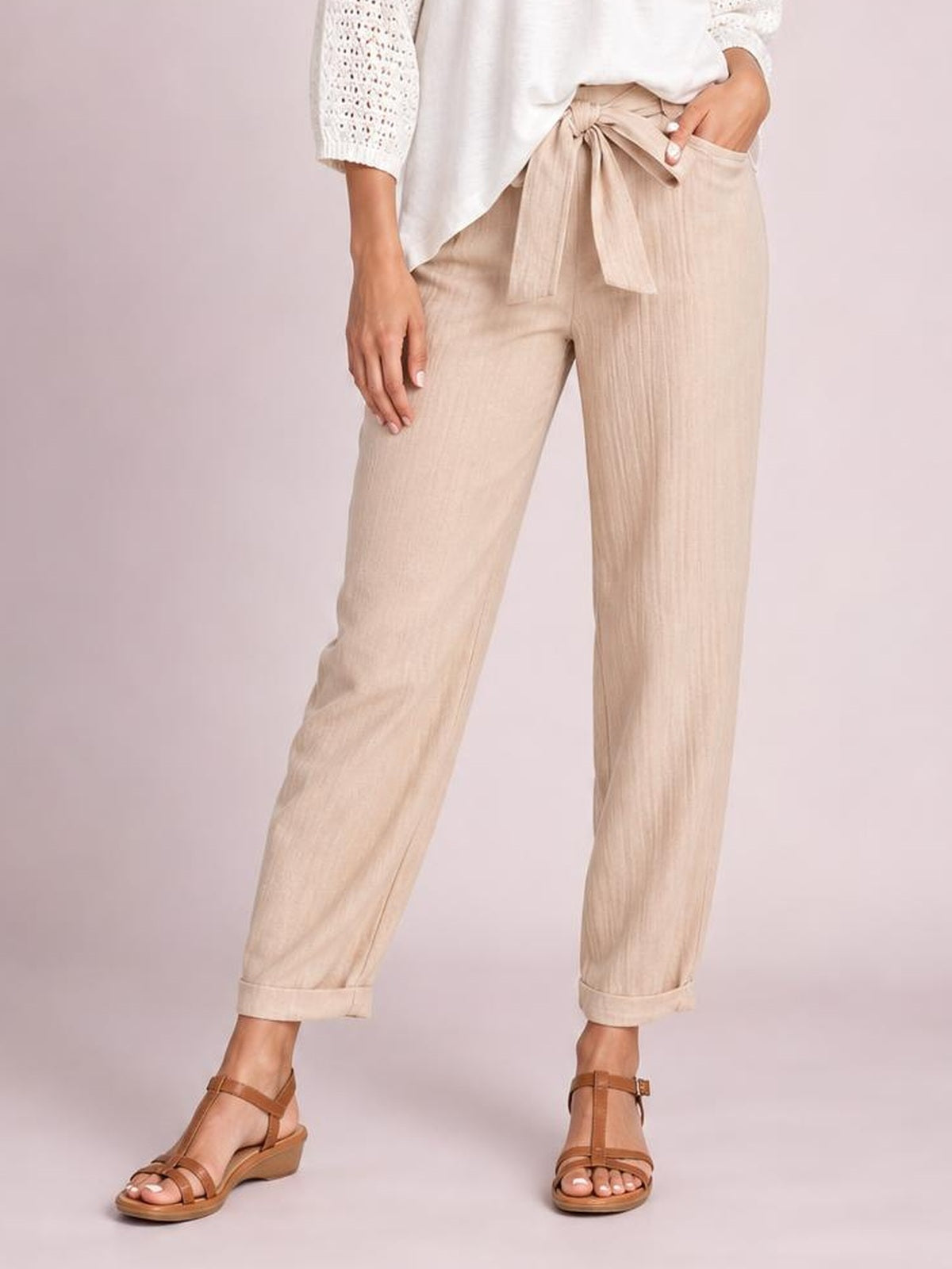 A model wears beige tied high-waist pants with a relaxed tapered silhouette, front pockets, and cuffed cropped hems while standing with one hand in a pocket.