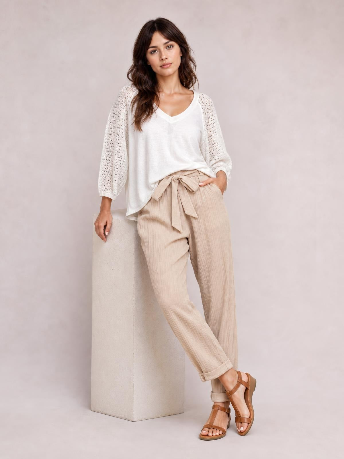Model poses beside a pedestal in beige tied high waist pants with a relaxed tapered silhouette, visible side pocket, textured fabric, and rolled cuffs.