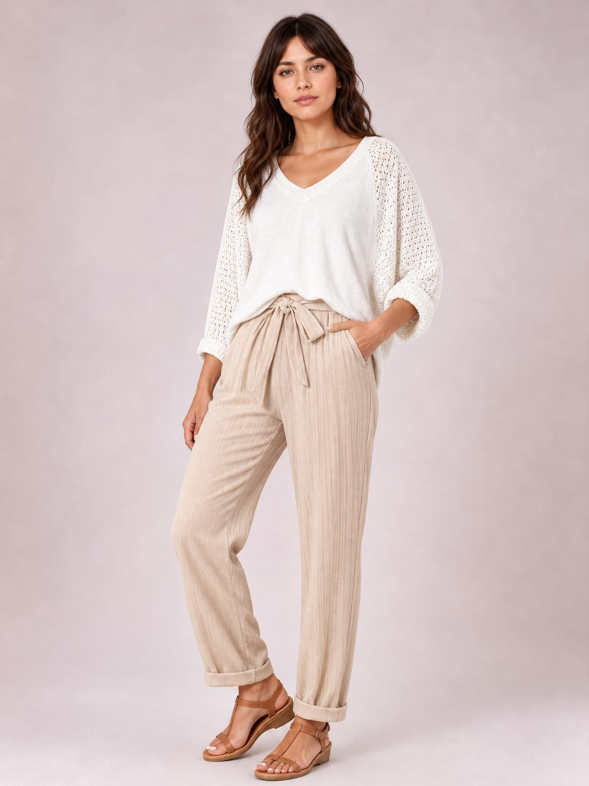 Model poses in beige tied high waist pants with button details, side pockets, a relaxed straight-leg silhouette, and rolled cuffs.