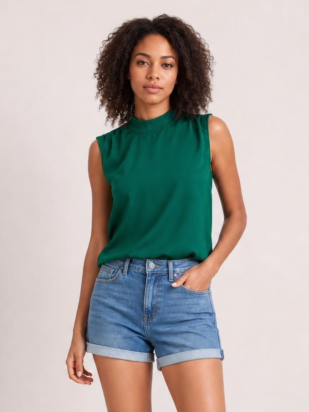 Model wearing a solid emerald green sleeveless blouse with a mock neck, gathered shoulder detailing, and a relaxed regular-length silhouette.