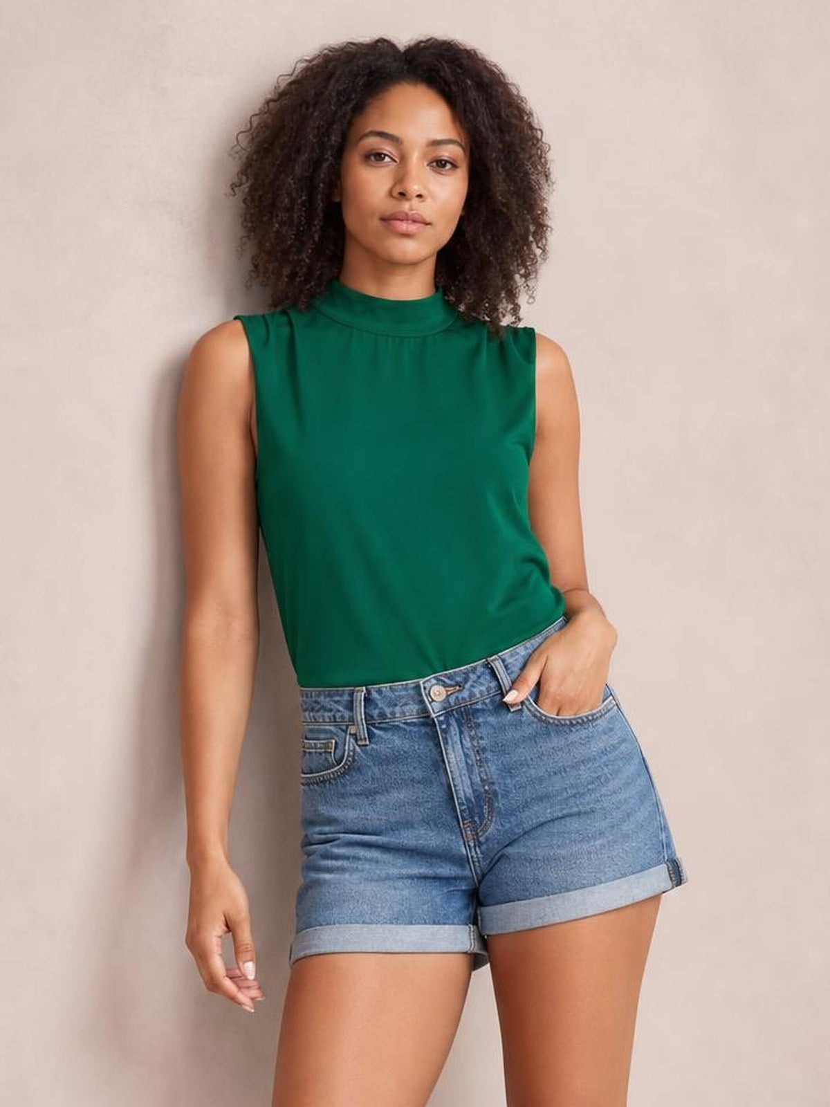 Model wears a solid green sleeveless blouse with a mock neck and relaxed regular-length silhouette, posing with one hand in her shorts pocket.
