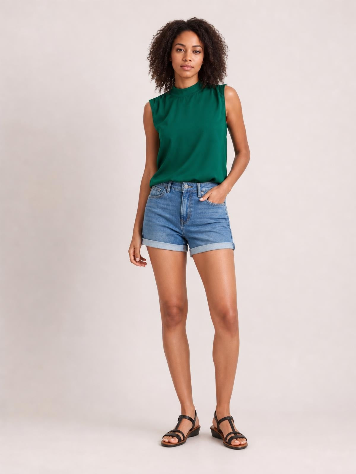 A model stands with one hand in her pocket wearing a solid emerald-green sleeveless blouse with a gathered mock neck and relaxed regular-length silhouette.