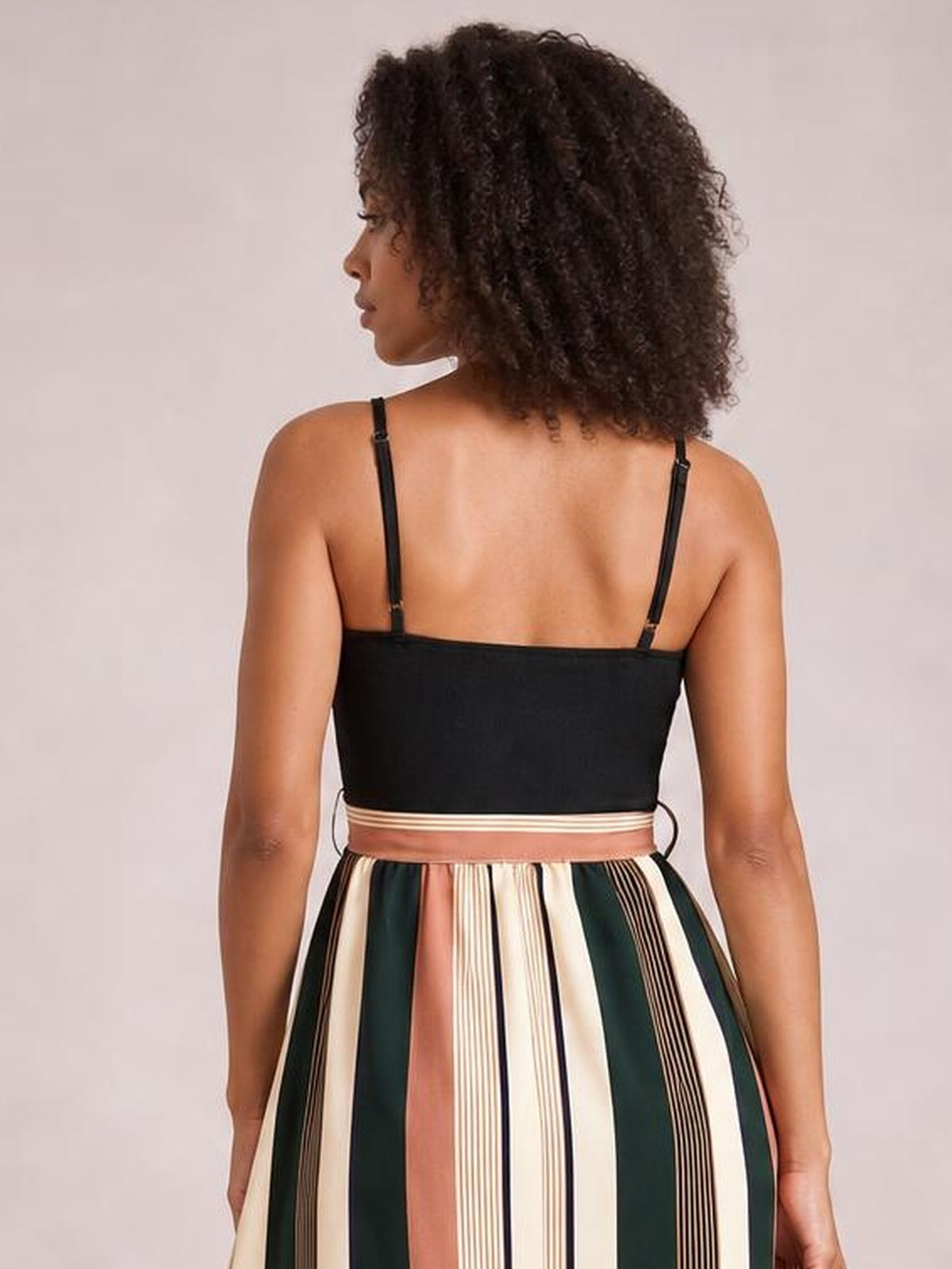 A model poses facing away in a striped spaghetti strap dress with a black fitted bodice, adjustable straps, waist ties, and a flowing multicolor vertical-striped skirt.