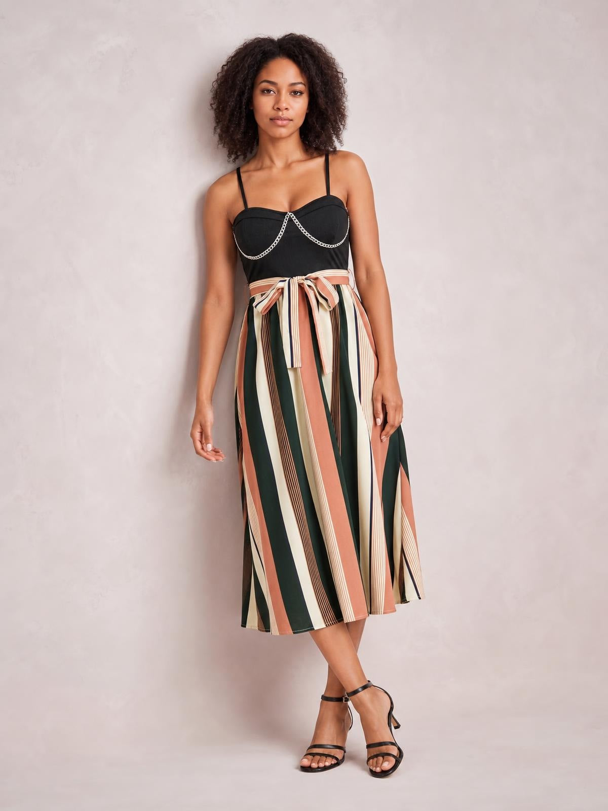Model stands with crossed legs in a tied striped spaghetti strap dress featuring a black fitted bodice and flowing vertical-striped midi skirt.