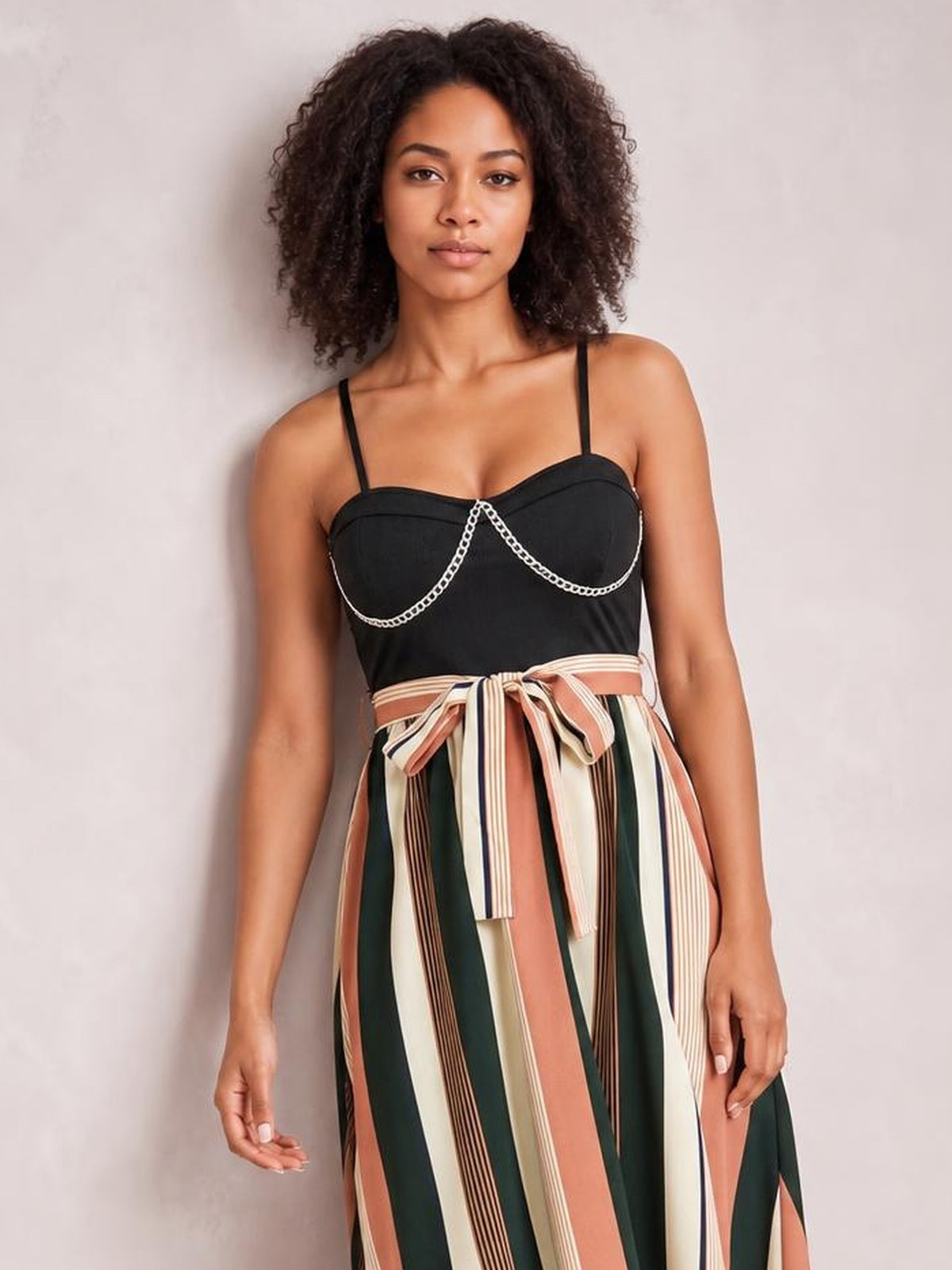 A model stands in a striped spaghetti strap dress with a black sweetheart bodice, vertical multicolor skirt, and tied waist bow.