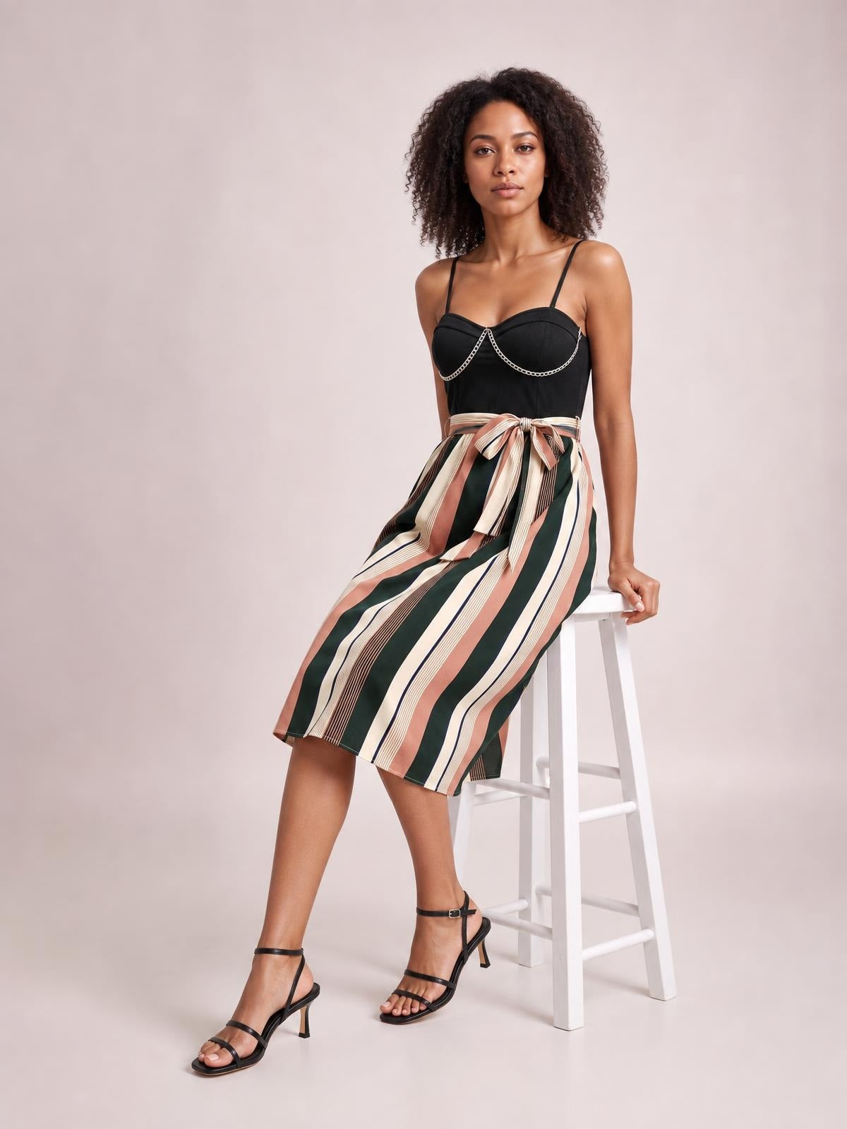 Model seated on a stool wears a tied striped spaghetti strap dress with a fitted black bustier bodice and flowing knee-length skirt in vertical stripes.