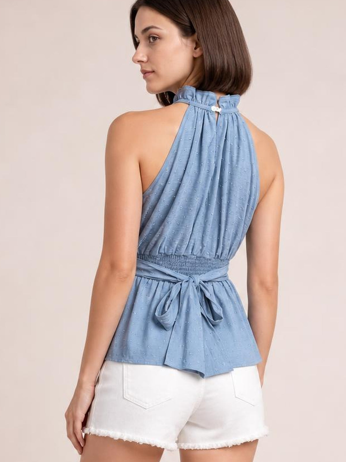A woman models a light blue Swiss dot mock neck tank from the back, featuring a sleeveless silhouette, gathered fabric, and a tied waist.