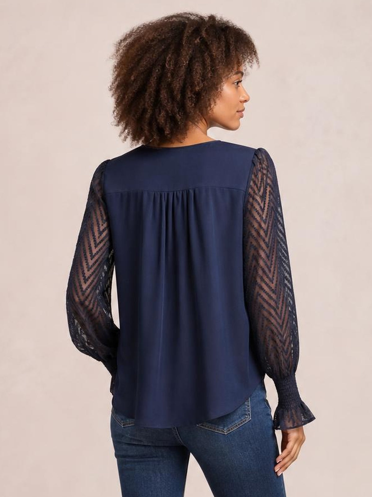 Model shown from the back wears a navy V-neck top with long patterned mesh sleeves, gathered back yoke, relaxed silhouette, flared cuffs, and curved hem.