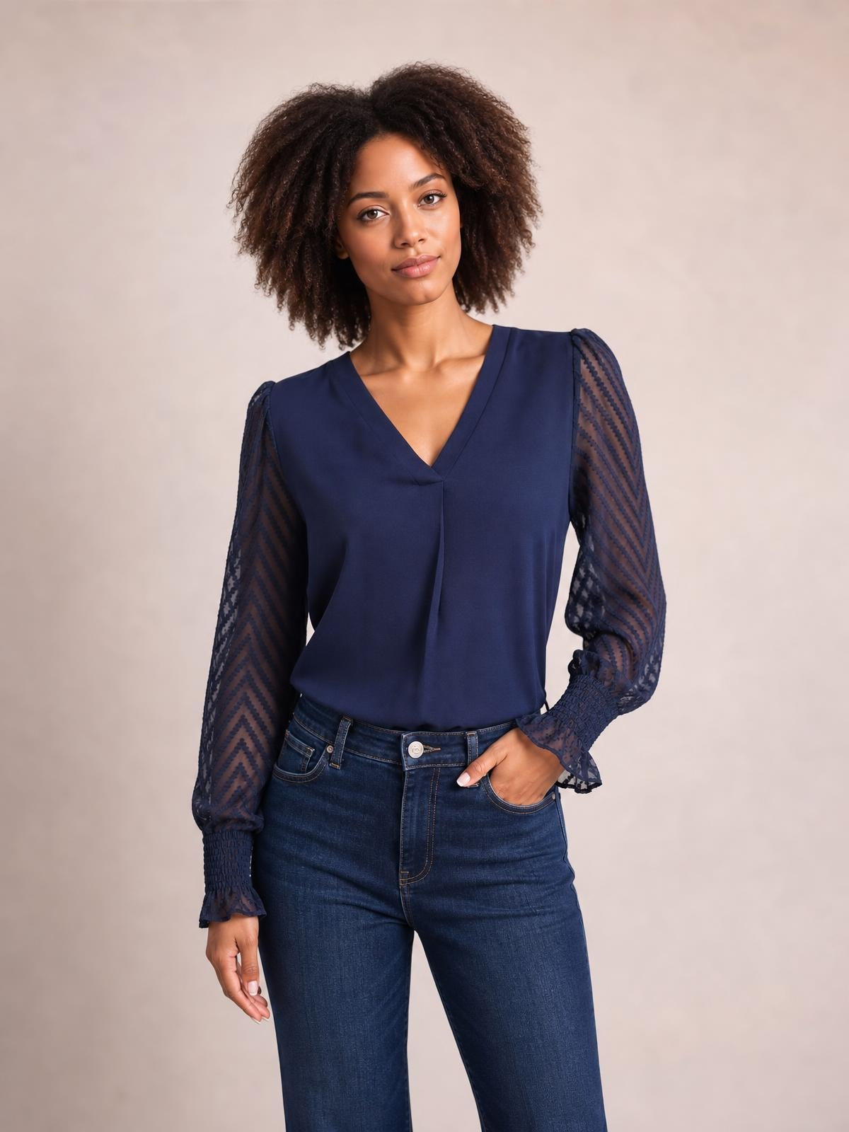 A model poses in a navy V-neck top with long sheer mesh sleeves, chevron texture, gathered cuffs, and a relaxed straight silhouette tucked into jeans.
