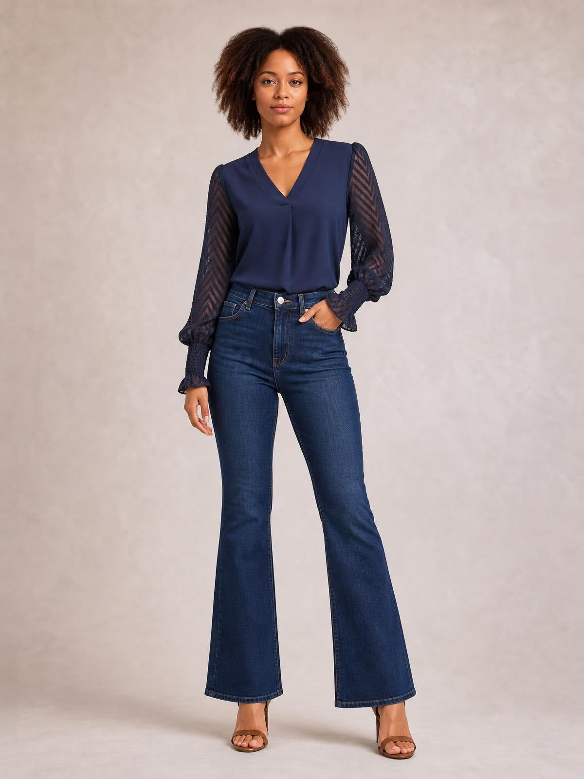 Model wearing a navy V-neck top with an opaque bodice, long sheer mesh sleeves, gathered cuffs, and a relaxed silhouette, posing with one hand in her jeans pocket.