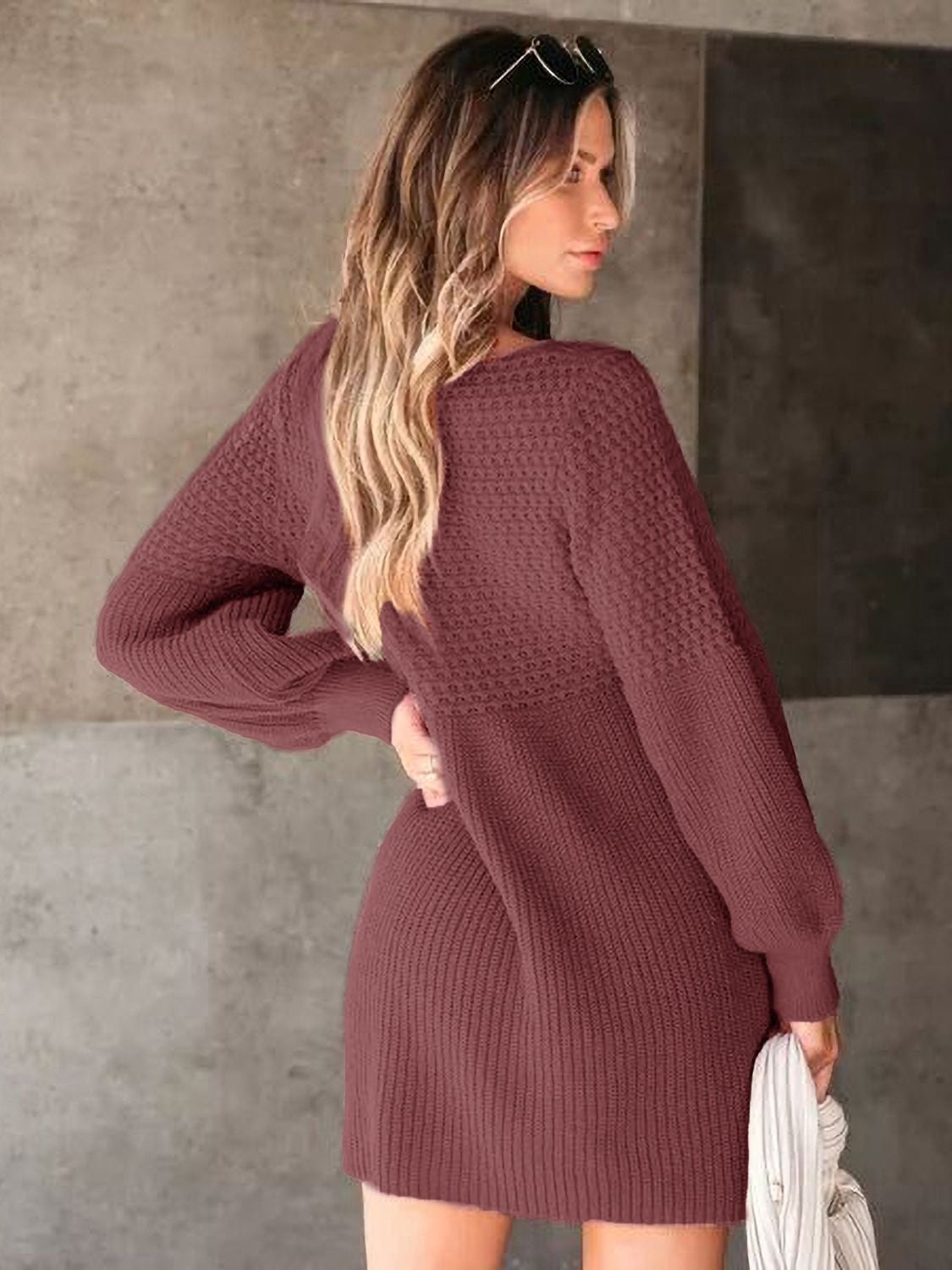 A model poses in a burgundy textured knit sweater dress with long sleeves, a relaxed above-knee silhouette, ribbed cuffs and hem, and one hand on her hip.