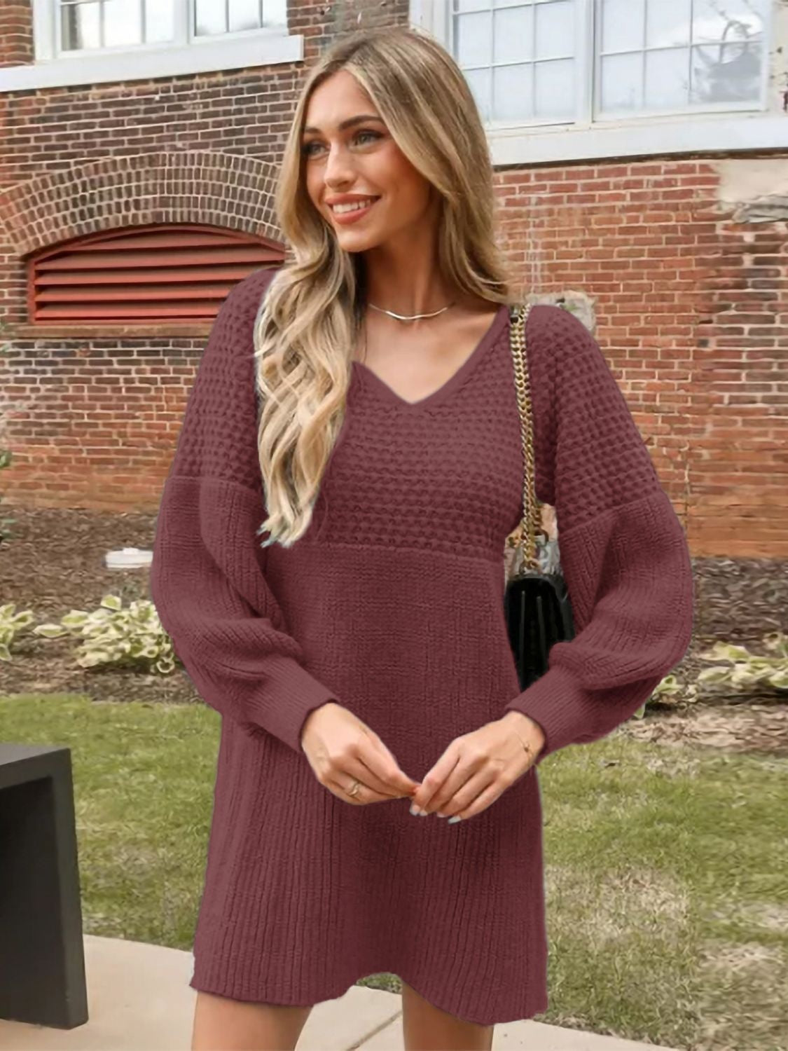 Model poses with hands clasped in a burgundy V-neck sweater dress featuring long sleeves, textured knit construction, and a relaxed above-knee silhouette.