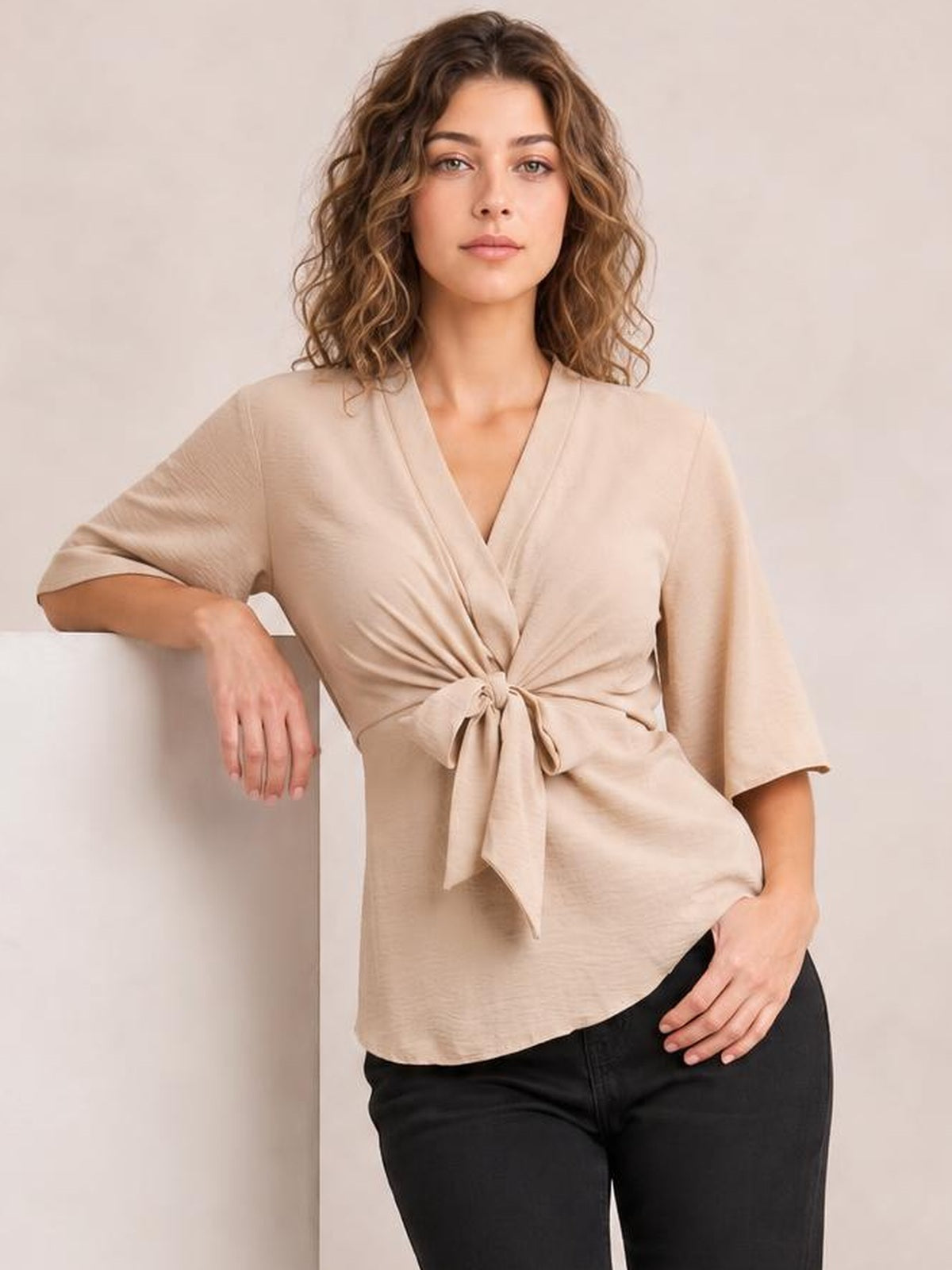 Model wearing a solid beige V-neck tie-front blouse with twisted waist detail, half-length regular sleeves, and a long relaxed silhouette.