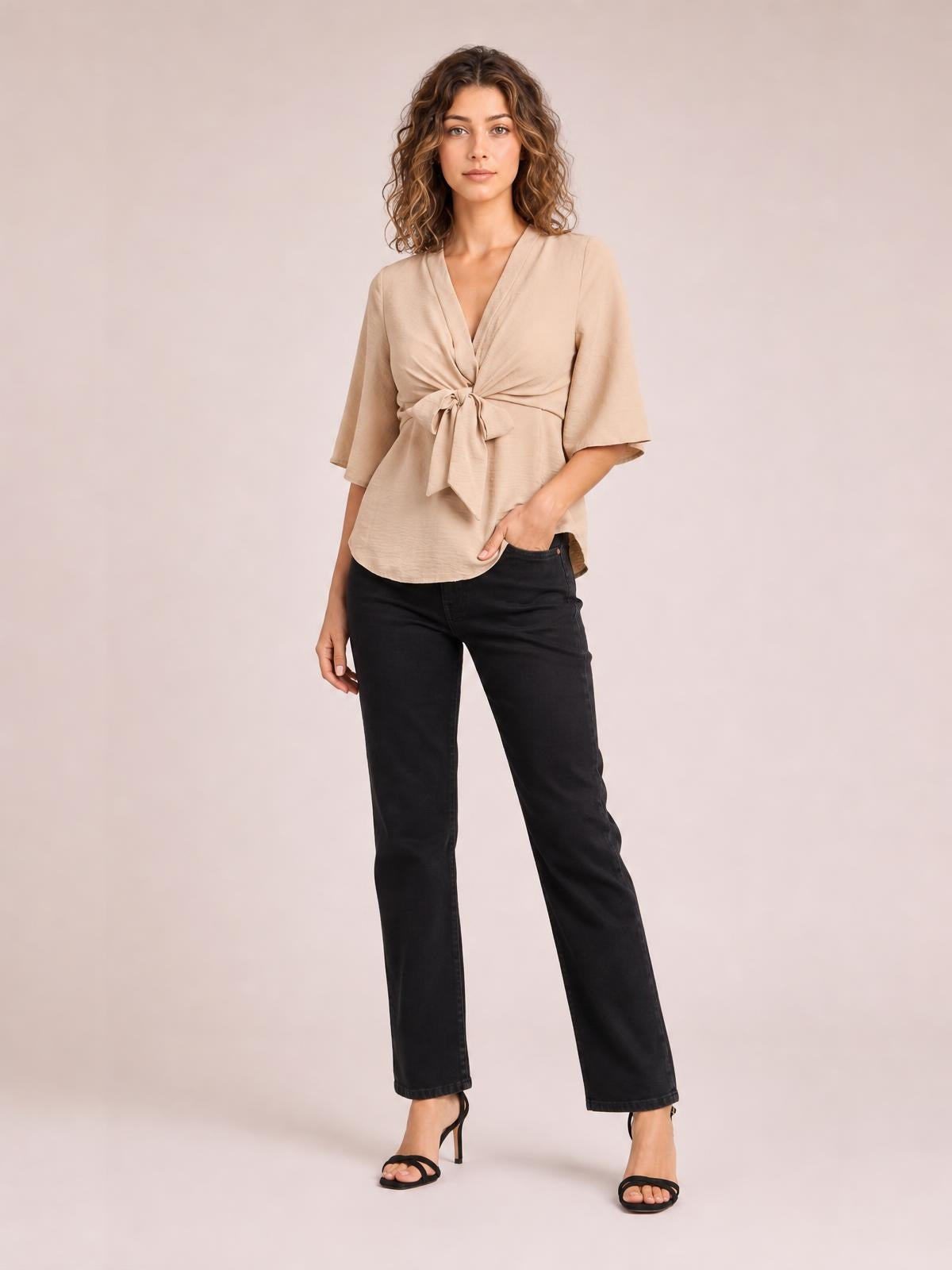 Model poses in a solid beige V-neck blouse with twisted tie-front detail, flowing half sleeves, and a long relaxed silhouette.